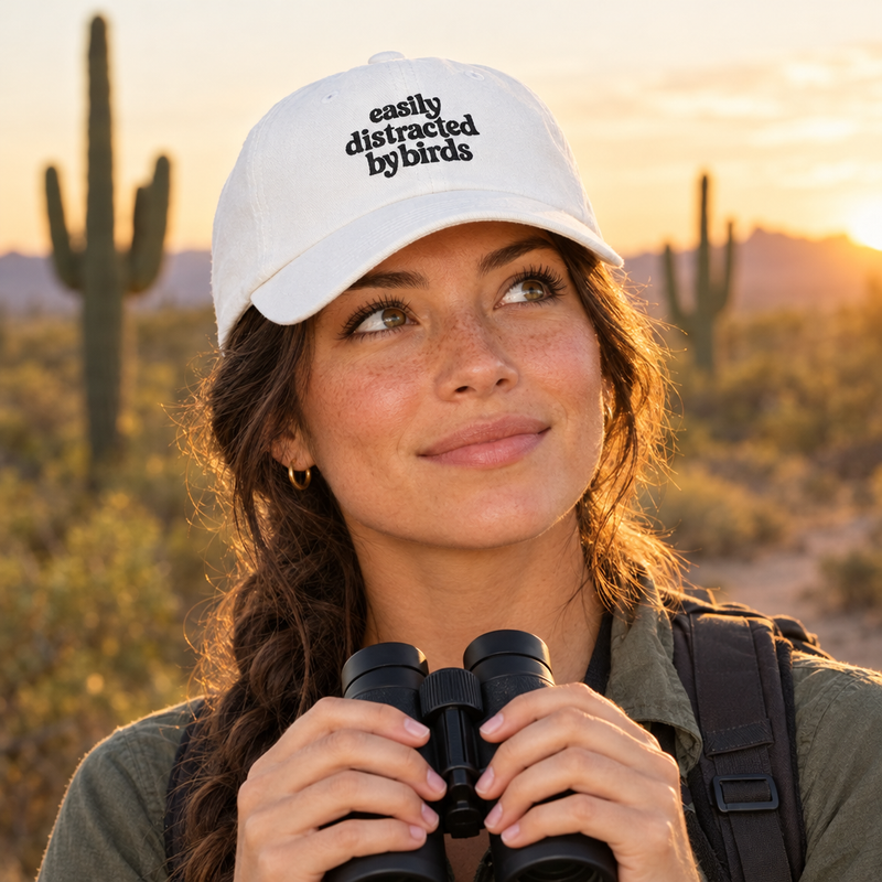 Woman on a desert trail wearing the white Easily Distracted by Birds dad cap, holding binoculars